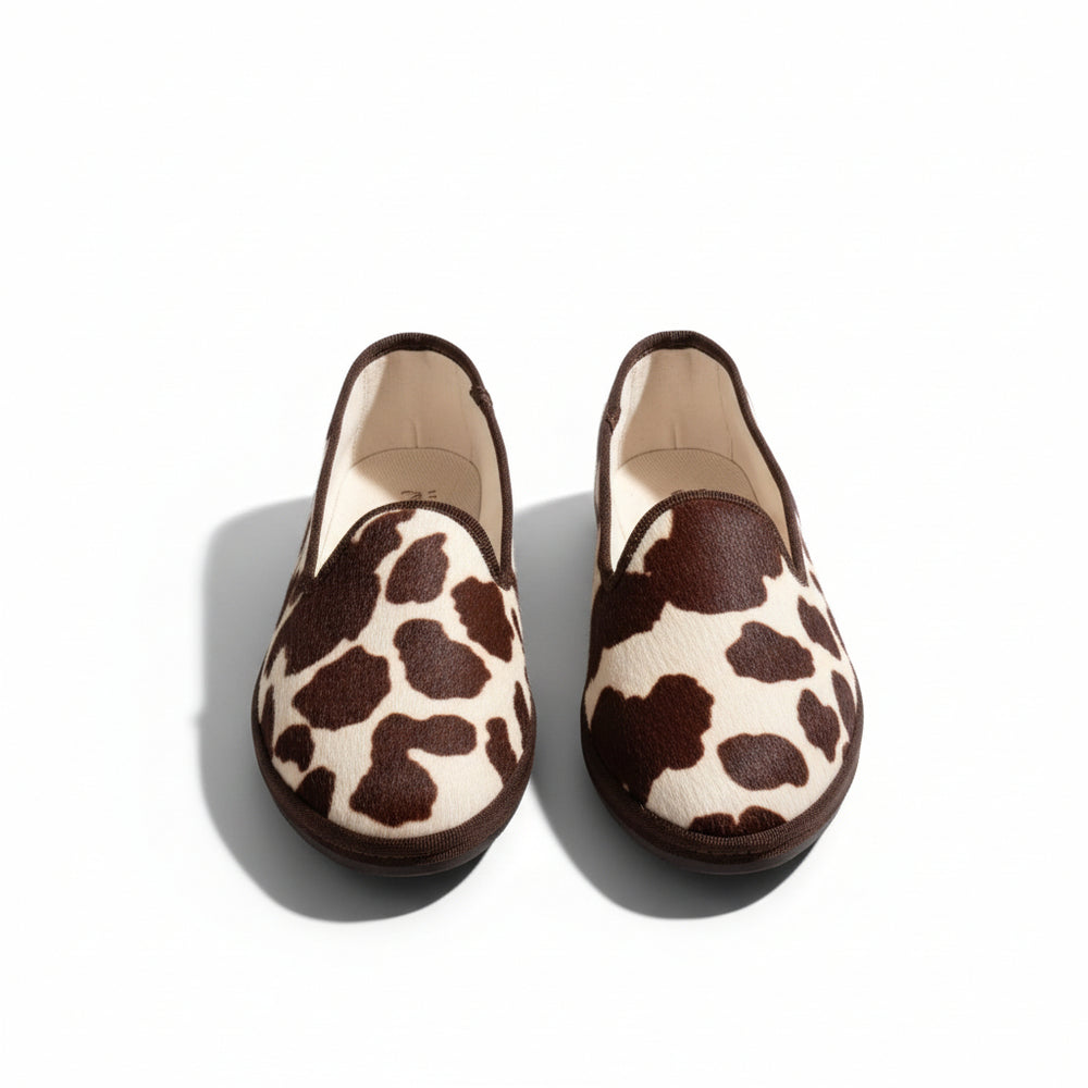 A pair of slip-on loafers with a brown and beige cow-print pattern, displayed on a dark tiled floor.