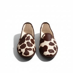 A pair of slip-on loafers with a brown and beige cow-print pattern, displayed on a dark tiled floor.