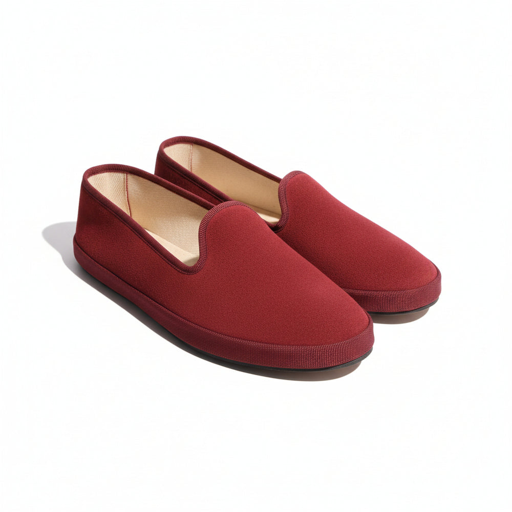 A pair of red suede slip-on slippers with a light beige interior lining, placed on a textured green marble surface.